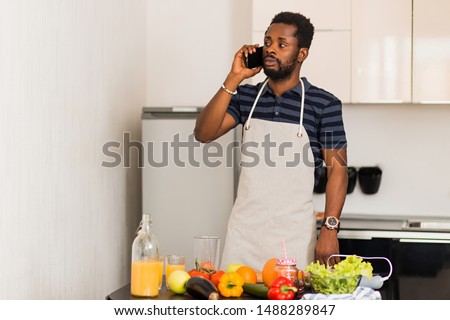 Handsome black guy wearing apron talking on the mobile phone while cooking, has thoughtful expression, does not know what to prepare, standing in kitchen at home.