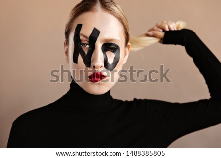 Woman with red lips and word no on face winks. Portrait of girl in black outfit holding her blond hair