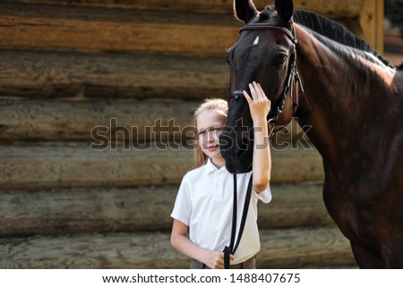 A teenage girl jockey stands next to a brown horse and hugs her. Against the background of a wooden wall stables on a green field.