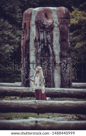 Mysterious young woman in folk costume and with panpipe in hand stays near old big wooden statue in autumn forest. Scary theme