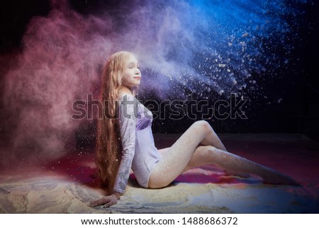 Beautiful teen girl with long blonde curly hair in a dark room with colored lights and clouds of flour. Sports teenager young model during a photoshoot with flying flour and color light