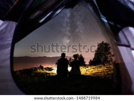 View from inside tent of young couple tourists having a rest at campfire near tent. Back view of man and woman enjoying night sky full of stars and Milky way. On the background starry sky, mountains