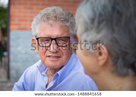 Portrait of an older man with grey hair and glasses, smiling at his wife in outdoor scenery. The woman is out of focus.