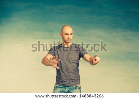 A young bald man in a gray T-shirt on a green background surprised looking at something examines, pushes and points to an invisible screen. 