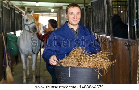 Man in working clothes feeding horse with hay at stable