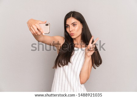 Image of charming brunette woman wearing dress showing peace sign while taking selfie photo on smartphone isolated over gray background