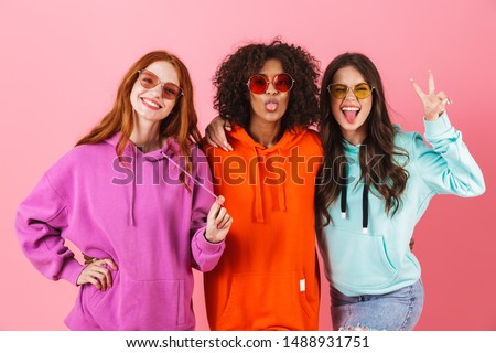 Three happy young girls wearing colorful hoodies standing isolated over pink background, looking at camera