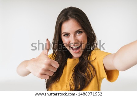 Image of excited brunette woman smiling and showing thumb up while taking selfie photo isolated over white background