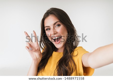 Image of pleased brunette woman smiling and showing peace sign while taking selfie photo isolated over white background