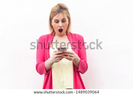 Portrait of shocked beautiful blogger young woman in pink blouse standing, use smartphone and watching video with amazed face, opened mouth. indoor, studio shot, isolated on white background