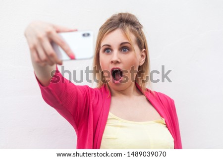 What?! Closeup portrait of unexpectedly attractive blogger woman in pink blouse standing, holding phone and making videocall with amazing face. indoor, studio shot, isolated on white background