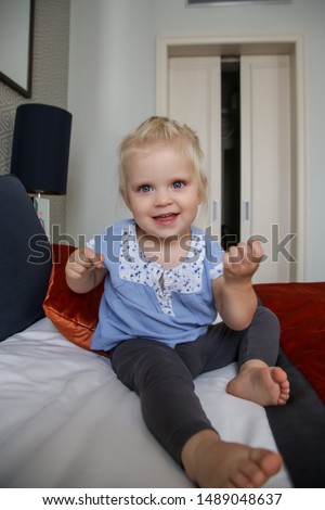 Cute blonde toddler girl in blue cotton shirt sitting on the bed 