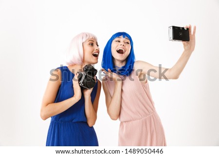 Portrait of two cheerful women wearing blue and pink wigs taking selfie photo on retro camera isolated over white background