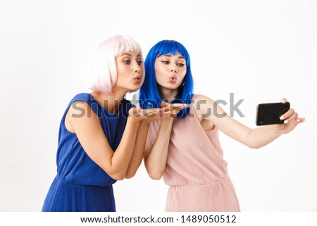 Portrait of two young women wearing blue and pink wigs taking selfie photo on cellphone and sending air kiss isolated over white background