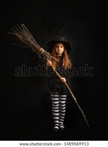 little witch girl in traditional Halloween dress and pointed hat is standing with a wooden whisk on a dark background