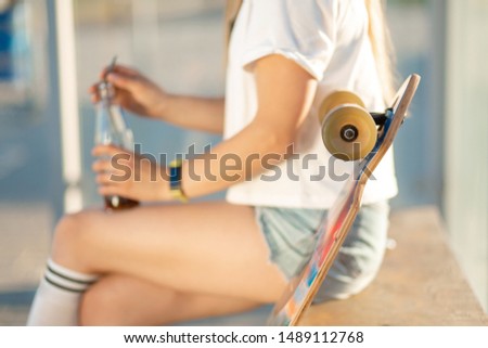 Stylish young girl sitting on bus stop with her longboard, drink soda and wait for bus.