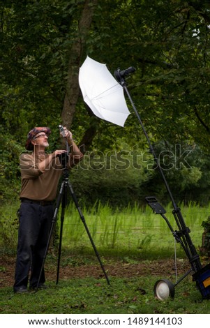 Mature male photographer gray beard glasses floppy hat brown shirt outside in green wooded area with DSLR digital camera on tripod testing umbrella strobe flash speedlite and flash exposure meter