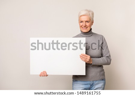 Mature woman with blank advertising board over light studio background, free space
