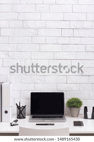 Modern white office desk with blank screen on laptop and supplies in front of bricks wall, vertical panorama, copy space