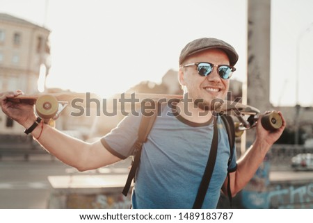 Happy hipster guy posing with skateboard in sunset in summer evening. Portrait of handsome stylish man model in blue shirt, sunglasses and hat on city street background