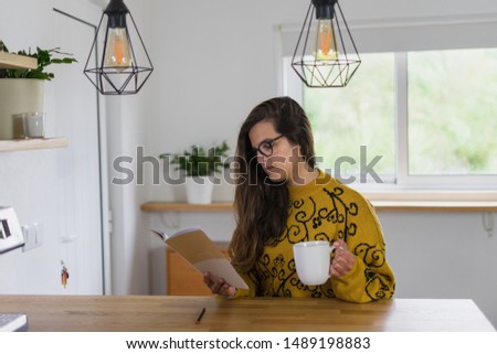 Woman with glasses holding a mug and reading from a notebook on a wooden workbench inside a house.