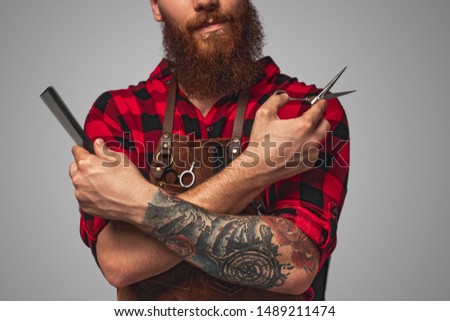 Unrecognizable bearded man in apron crossing tattooed arms with comb and scissors while working in salon against gray background