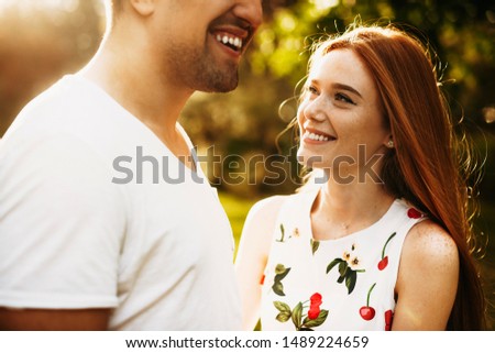 Side view portrait of a lovely young female with freckles and long red hair smiling while looking at her boyfriend outside against sunrise and trees.