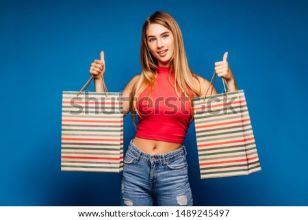 Pretty elegant woman in a red t-shirt and denim jeans with packages shopping blue background. Okay sign.