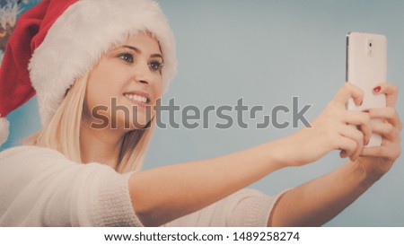 Enjoying christmas gifts. Woman in santa hat taking picture of herself using smart phone. Indoor shot on blue background