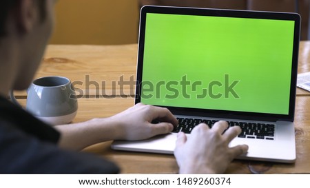 View from the back of man in black shirt sitting at the table and typing on laptop with green chroma key screen. Stock footage. Laptop new technology concept, chroma key green screen