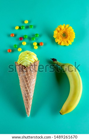 yellow marshmallows in ice cream cone with banana, yellow gerbera flower and colorful round candies on aquamarine background