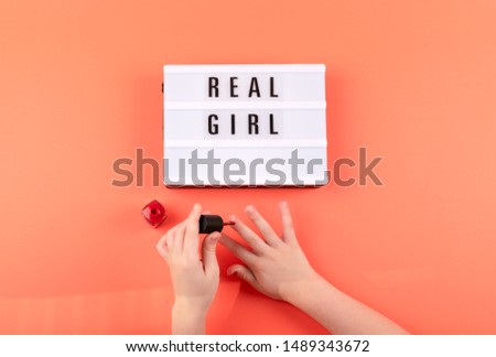 Real girl text on lightbox and girl hands paints nails with red nail polish on coral background. Beauty, makeup flat lay