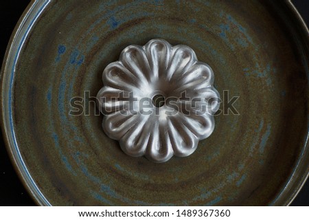A decorative, stamped from metal flower of silver color lies on a textured, swamp color, ceramic plate close-up.
