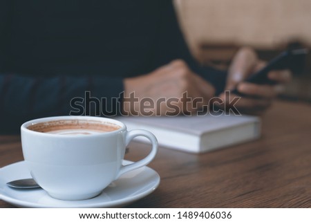 Closeup image of casual business man using mobile smart phone connecting free wifi browsing internet with book, latte coffee on wooden table in coffee shop, vintage style.