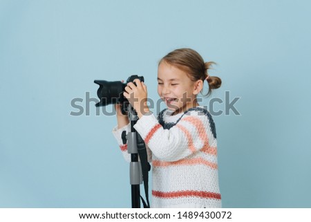 Little first grader girl laughing with her eyes closed, about to take a picture with the camera on tripod. She has low double buns, wearing knitted sweater and jeans shorts. Over blue background.