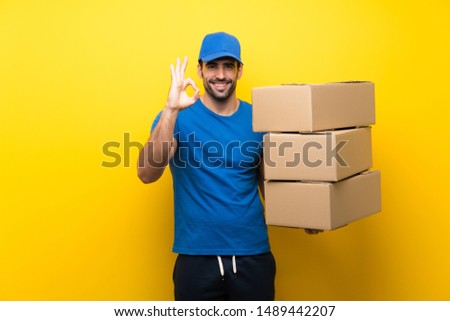 Young delivery man over isolated yellow wall showing ok sign with fingers