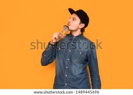 A young guy in a black cap and in a denim shirt drinks water from a glass from the heat. The guy stands on an orange background and drinks water from thirst. Isolated in orange background.