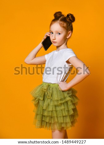 Beautiful red-haired girl child in a white T-shirt and a green skirt tutu is seriously talking on a mobile phone
