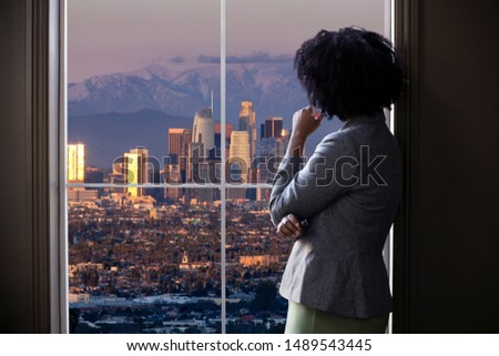 Black female business woman looking out the window of an office in Los Angeles.  She looks like a female architect thinking of urban development or a city mayor or governor planning zoning laws. 