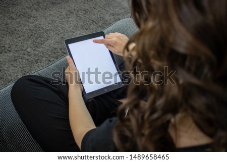 Young brunette woman playing on a tablet sat on a sofa relaxing, the tablet has a white full size screen to enable adding text or images. 