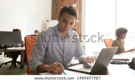 Focused male worker sit at desk in coworking space check data in document working at laptop, concentrated man employee analyzing paperwork busy typing at computer in shared office