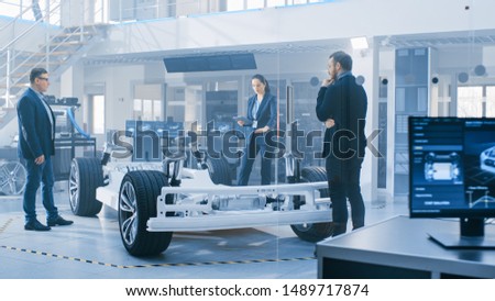 Automotive Design Engineers Talking while Working on Electric Car Chassis Prototype. In Innovation Laboratory Facility Concept Vehicle Frame Includes Wheels, Suspension, Engine and Battery.