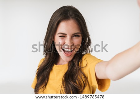 Image of happy brunette woman wearing casual t-shirt smiling and looking at camera while taking selfie photo isolated over white background