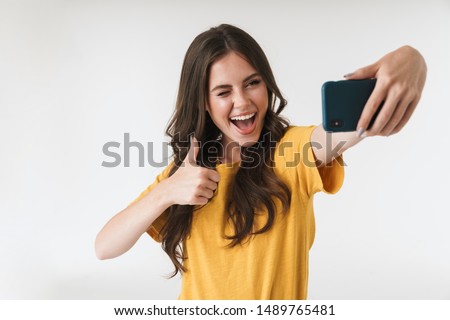 Image of young brunette woman laughing and showing thumb up while taking selfie photo on cellphone isolated over white background