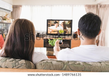 Asian male and female couples are watching TV on the sofa in the house. Royalty-Free Stock Photo #1489766135