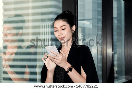 Happy Asian woman in suit using smartphone standing in modern office beside glass wall with office buildings in the background.Executive businesswoman working with a mobile phone in the office.