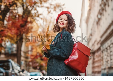 Outdoor autumn portrait of young fashionable happy smiling curly girl wearing gray coat, beret, with red leather backpack, posing in street of European city. Copy, empty space for text 