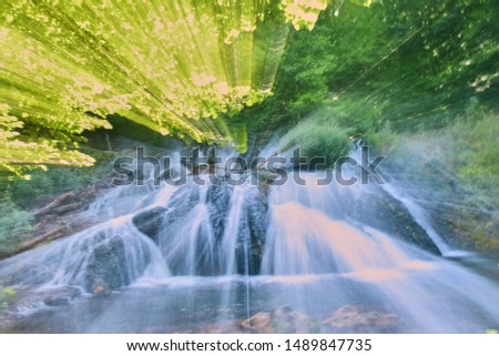 Photo of the waterfall, taken at a long shutter speed with a change in focal length. Abstract drawing.Blurred water flow, sunlight, lots of greenery around.