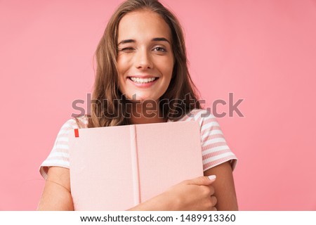 Image closeup of content pretty woman smiling at camera while hugging her diary book isolated over pink background