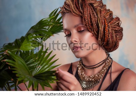 Southern flavor. A young tanned woman with afro braids smiles holding a big pot with a tropical plant. On her neck she has large jewels in ethnic style.